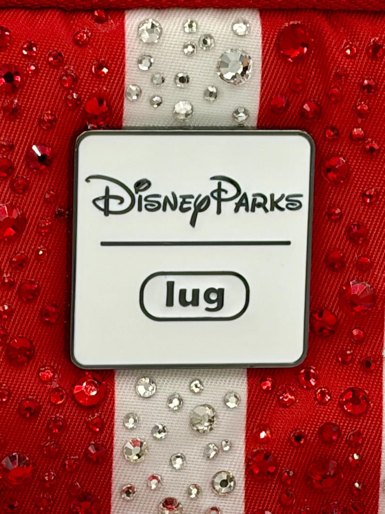 May include: A rectangular white metal plaque with rounded corners featuring the black text Disney Parks and the word lug inside a black-outlined oval. The plaque is centered on a red fabric background with vertical white stripes adorned with clear crystals.