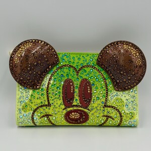 May include: A lime green wallet featuring a bedazzled Mickey Mouse face. The ears are brown with gold accents, and the face is decorated with green, brown, and clear rhinestones. The wallet has a zipper closure and a light-colored zipper pull.