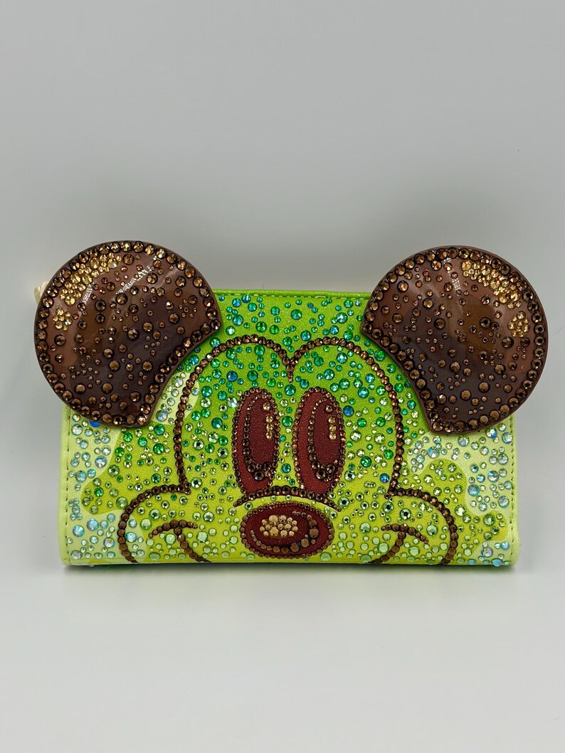 May include: A lime green wallet featuring a bedazzled Mickey Mouse face. The ears are brown with gold accents, and the face is decorated with green, brown, and clear rhinestones. The wallet has a zipper closure and a light-colored zipper pull.