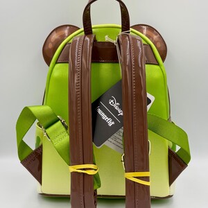 May include: A green and brown backpack with a cartoon character design. The backpack has brown straps, a top handle, and ear-shaped accents. The straps are secured with yellow rubber bands. A tag with the "Loungefly" and "Disney" logos is attached.