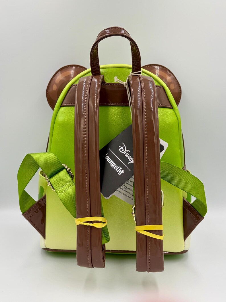 May include: A green and brown backpack with a cartoon character design. The backpack has brown straps, a top handle, and ear-shaped accents. The straps are secured with yellow rubber bands. A tag with the "Loungefly" and "Disney" logos is attached.