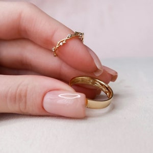 May include: Two gold rings on a finger. The top ring is a delicate band with a vine design and small diamonds. The bottom ring is a simple, smooth band.