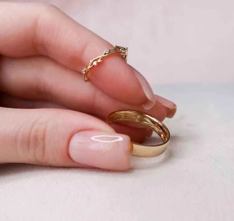 May include: Two gold rings on a finger. The top ring is a delicate band with a vine design and small diamonds. The bottom ring is a simple, smooth band.