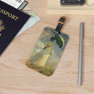 May include: Luggage tag featuring a painting of a woman with a parasol in a field. The tag has a black leather strap and a gold-colored buckle. A silver pen and a passport are also visible.