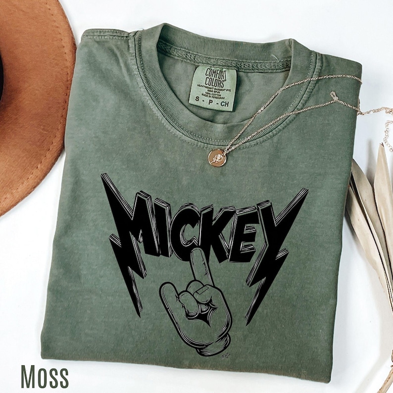 Op de afbeelding: Een mosgroen t-shirt met een zwarte afbeelding van "MICKEY" met een hand die de middelvinger opsteekt. Het shirt heeft een ronde hals en korte mouwen. Een gouden ketting met een hanger is over het shirt gedrapeerd.