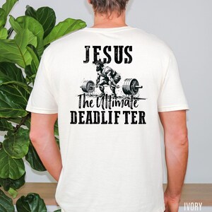 May include: Ivory-colored t-shirt featuring the text "JESUS" above an illustration of a weightlifter performing a deadlift. Below the illustration, the text reads "The Ultimate DEADLIFTER" in a bold, black font. The shirt is a short-sleeved crew neck.