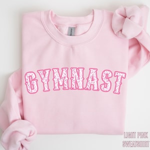 May include: A light pink sweatshirt with the word "GYMNAST" in a pink and white floral design across the front. The sweatshirt has a crew neck and long sleeves. The text "LIGHT PINK SWEATSHIRT" is at the bottom right.