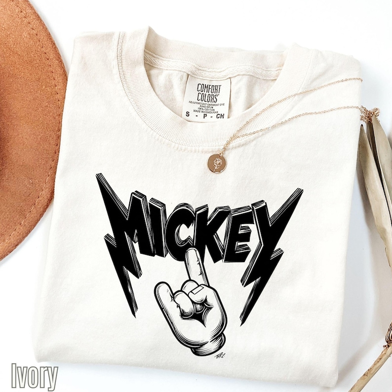 Op de afbeelding: Ivoor T-shirt met een zwarte "MICKEY" afbeelding en handgebaar. Het shirt heeft een ronde hals en een "COMFORT COLORS" label. Een gouden ketting met hanger is over het shirt gedrapeerd.