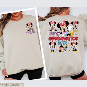 A woman wearing a beige sweatshirt with a design featuring Minnie Mouse and the text "In my gymatics era" printed on the back.