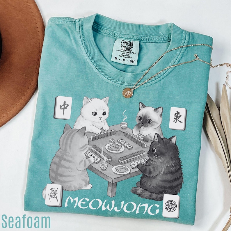 A turquoise t-shirt with a cat-themed graphic design, featuring four cats playing mahjong.