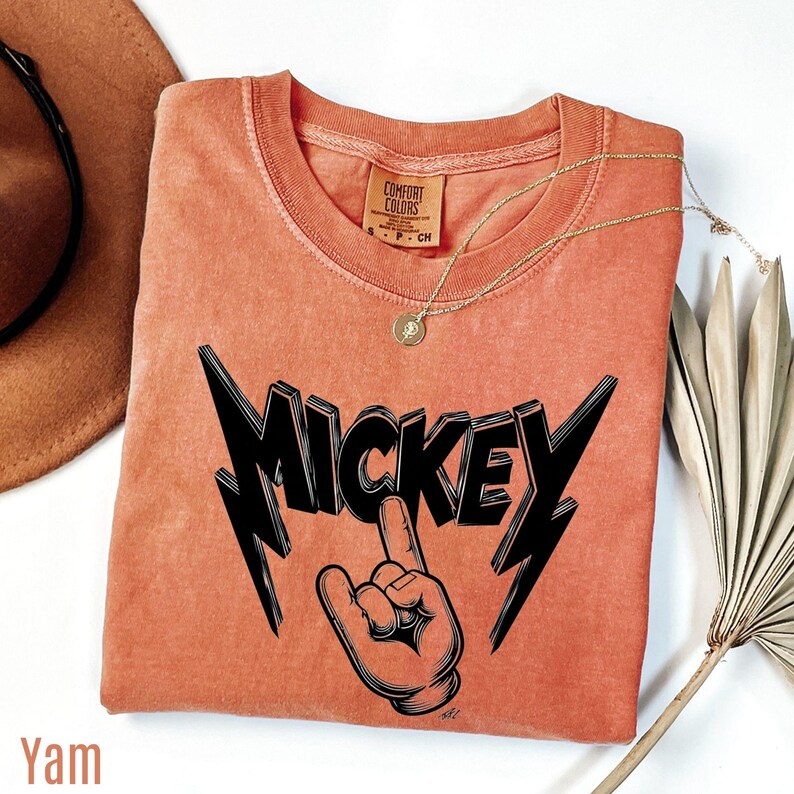 Op de afbeelding: Een koraalkleurig t-shirt met een zwarte afbeelding met de tekst "MICKEY" met een hand die een rock and roll-gebaar maakt. De shirt heeft een ronde hals en een klein label met de tekst "COMFORT COLORS". Een gouden ketting en een bruine hoed staan ook op de foto.