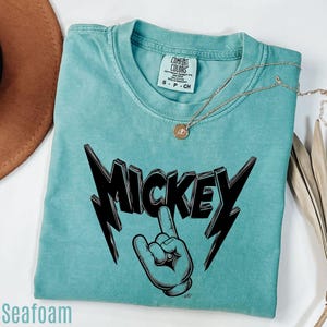 Op de afbeelding: Zeegroen t-shirt met een zwarte "MICKEY" afbeelding en handgebaar. Het shirt heeft een ronde hals en korte mouwen. Een gouden ketting met een hanger ligt op het shirt.