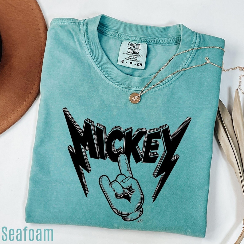 Op de afbeelding: Zeegroen t-shirt met een zwarte "MICKEY" afbeelding en handgebaar. Het shirt heeft een ronde hals en korte mouwen. Een gouden ketting met een hanger ligt op het shirt.