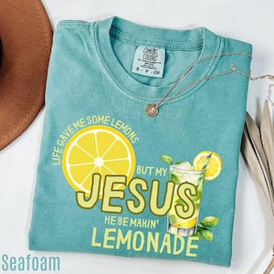 May include: Seafoam green t-shirt with the text "Life gave me some lemons but my Jesus he be makin' lemonade" and a graphic of a lemon and a glass of lemonade. A gold necklace is draped across the shirt.