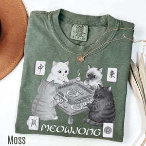 May include: Moss green t-shirt featuring a graphic of four cats playing Mahjong. The text "MEOWJONG" is printed below the table. A gold necklace is draped over the shirt. The shirt is labeled "Comfort Colors" and is size small.