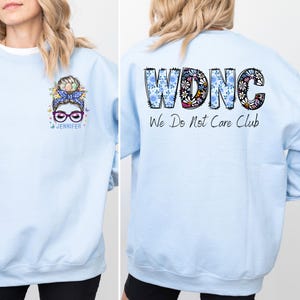 May include: Light blue sweatshirt with a graphic of a woman with glasses and a floral bun on the front. The back of the sweatshirt features the text "WDNC We Do Not Care Club" in a floral design. The sweatshirt is a casual style.