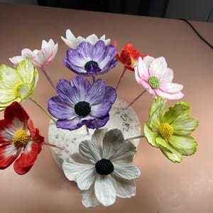 May include: A bouquet of paper flowers in various colors, including white, purple, pink, red, and green. The flowers are arranged in a circular foam base and are attached to wooden skewers.