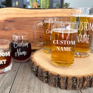 May include: Four glass beverage containers with custom engraved text. The containers include a large beer stein, two smaller beer glasses, and a rocks glass. The glasses are sitting on a wooden slice.