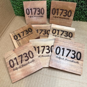 May include: A collection of wooden square coasters with the engraved postal code "01730" and the town name "Bedford, Massachusetts". The coasters are made of light brown wood and are arranged on a surface with green artificial grass.