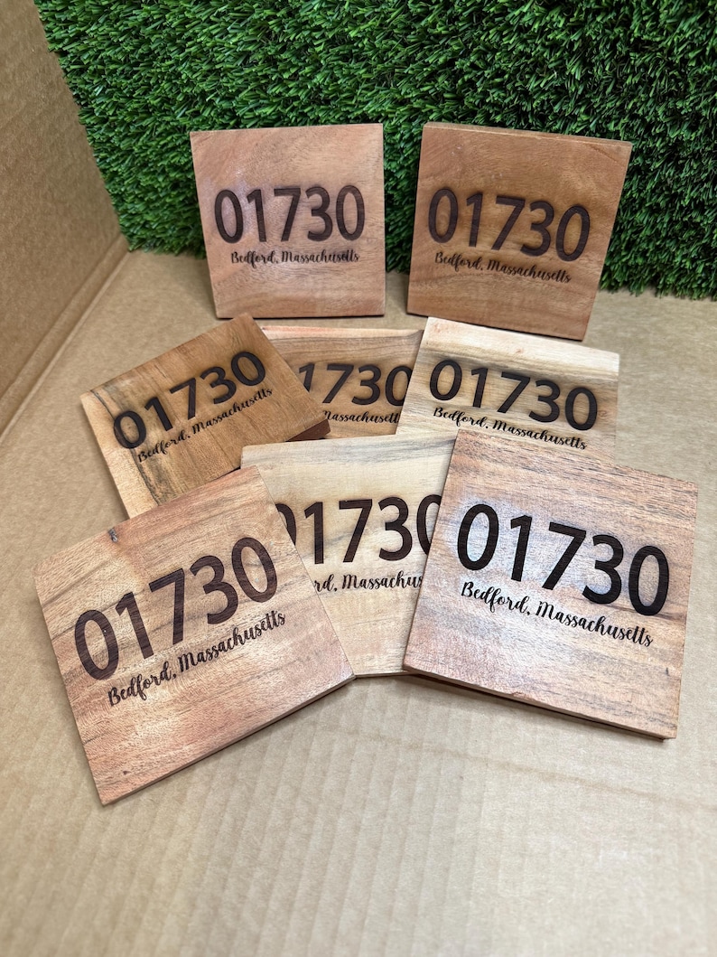 May include: A collection of wooden square coasters with the engraved postal code "01730" and the town name "Bedford, Massachusetts". The coasters are made of light brown wood and are arranged on a surface with green artificial grass.