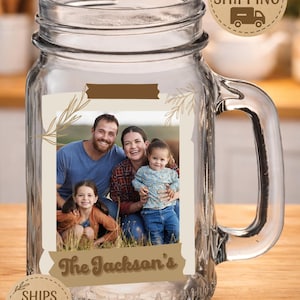 May include: A clear glass mason jar mug featuring a rectangular photo of a family of four. The photo is framed by a beige border with brown botanical line art. Below the photo, the text The Jackson's is printed in a brown script font.