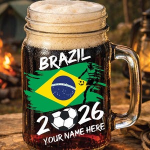 May include: A clear glass beer mug filled with dark liquid and foam. The mug features the word "BRAZIL" and a Brazilian flag design. A soccer player graphic and the numbers "2026" are also displayed. The text "YOUR NAME HERE" is at the bottom.