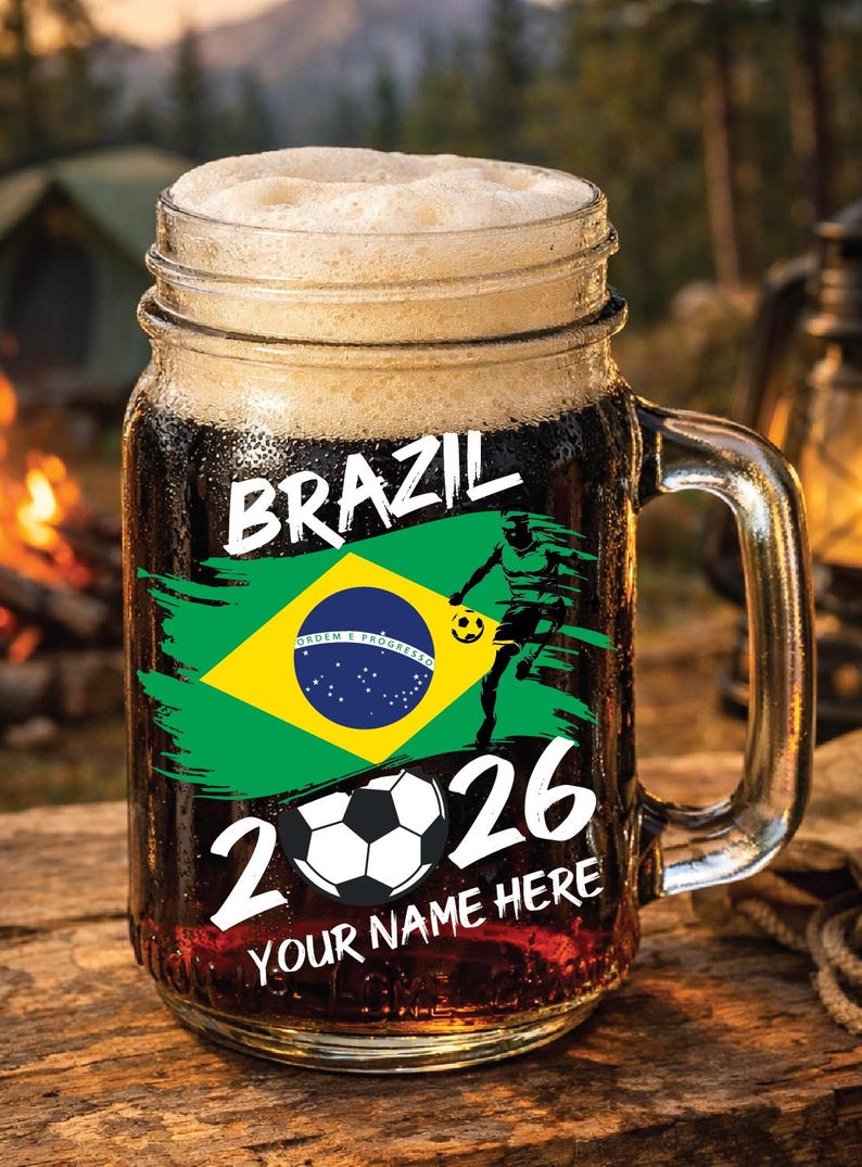 May include: A clear glass beer mug filled with dark liquid and foam. The mug features the word "BRAZIL" and a Brazilian flag design. A soccer player graphic and the numbers "2026" are also displayed. The text "YOUR NAME HERE" is at the bottom.