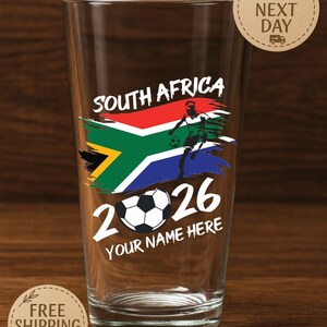 May include: A clear pint glass with a South Africa design. The design includes the South African flag, a soccer player, the year 2026, and the text "YOUR NAME HERE". The glass also has the text "SOUTH AFRICA".