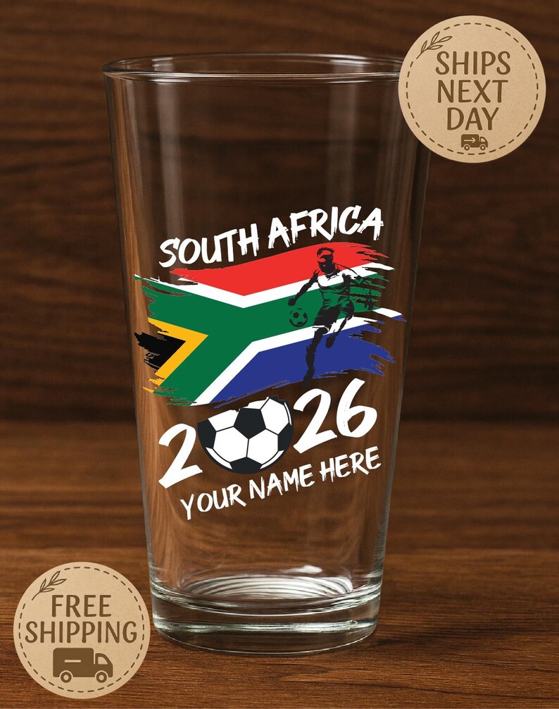 May include: A clear pint glass with a South Africa design. The design includes the South African flag, a soccer player, the year 2026, and the text "YOUR NAME HERE". The glass also has the text "SOUTH AFRICA".