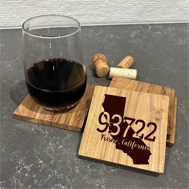 May include: A stemless wine glass filled with red wine rests on a wooden coaster. Additional coasters and wine corks are nearby. One coaster features the California outline with the zip code 93722 and "Fresno, California" text.