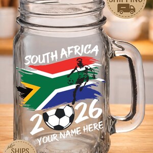 May include: Clear glass mason jar with a handle, featuring a South African flag design and a soccer player graphic. The text "SOUTH AFRICA 2026" and "YOUR NAME HERE" are also displayed. Includes a soccer ball graphic.