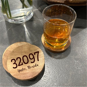 May include: Wooden coasters with the address 32097, Yulee, Florida, are displayed next to a glass of amber liquid. The coasters are round and made of light-colored wood. A glass of liquid sits on a matching coaster.