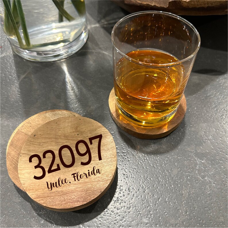 May include: Wooden coasters with the address 32097, Yulee, Florida, are displayed next to a glass of amber liquid. The coasters are round and made of light-colored wood. A glass of liquid sits on a matching coaster.