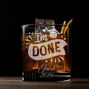 May include: A clear glass tumbler with a brown liquid and ice cubes. The glass has the text "I'm Done" with a graduation cap and stars. The text "Jacob 2023" is also on the glass.