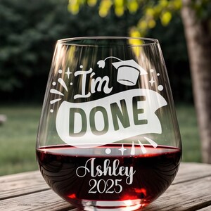 May include: A clear glass wine glass with the text "I'm Done" and a graduation cap etched on the side. The glass is filled with red wine and has the name "Ashley" and the year "2025" etched on the bottom.