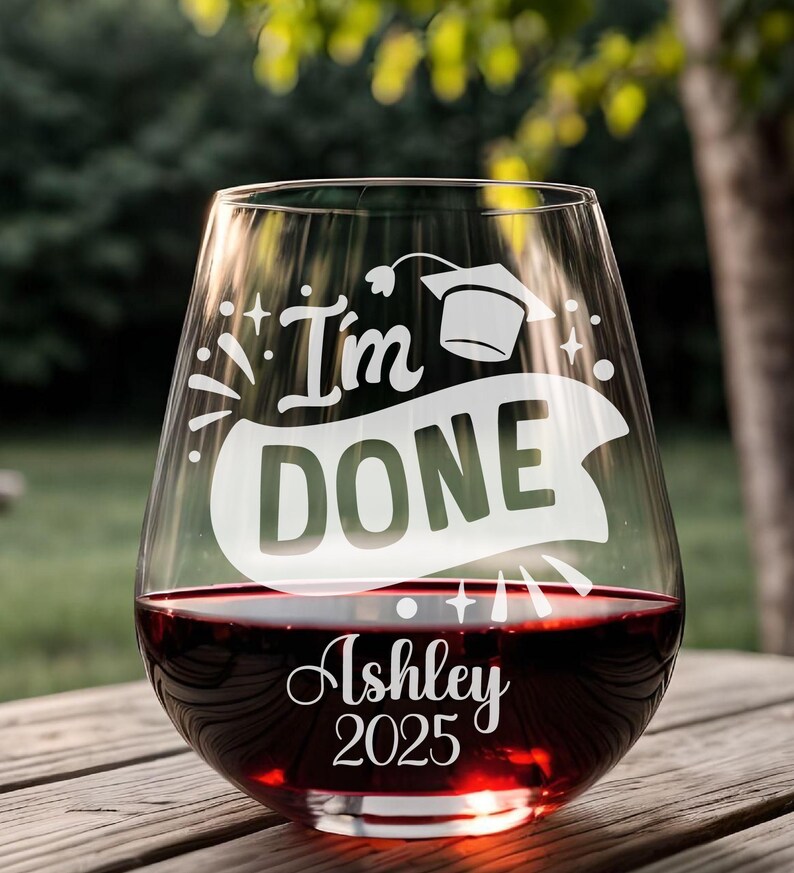 May include: A clear glass wine glass with the text "I'm Done" and a graduation cap etched on the side. The glass is filled with red wine and has the name "Ashley" and the year "2025" etched on the bottom.
