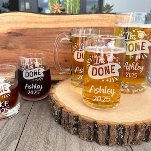 May include: Four glass beverage containers with the text "I'm Done" and a graduation cap graphic. The containers are on a wooden surface. The text "Ashley 2025" is on two of the containers. The text "Mike 2025" is on one of the containers.