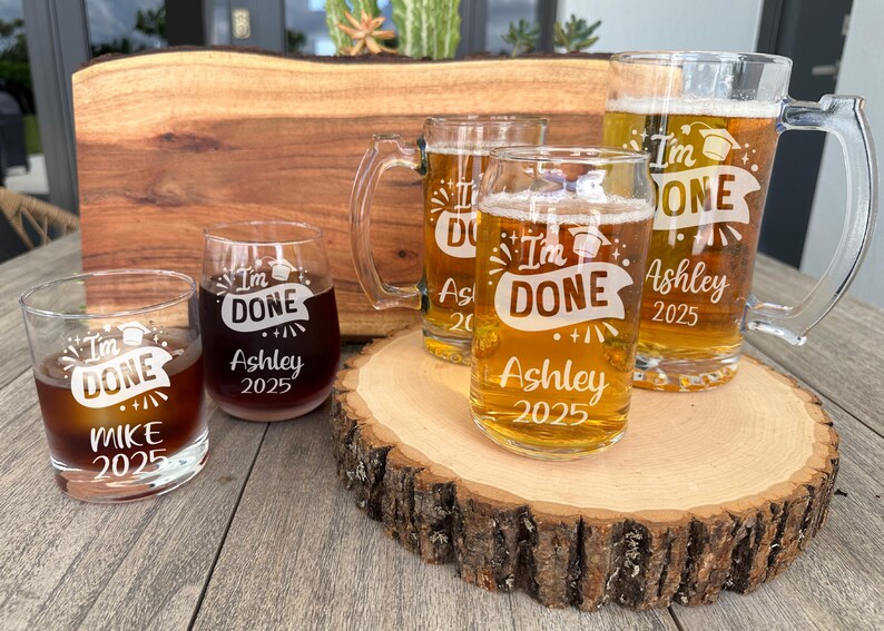 May include: Four glass beverage containers with the text "I'm Done" and a graduation cap graphic. The containers are on a wooden surface. The text "Ashley 2025" is on two of the containers. The text "Mike 2025" is on one of the containers.