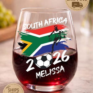 May include: Stemless wine glass filled with red liquid, featuring a South Africa flag design, a soccer player graphic, and the text "SOUTH AFRICA 2026 MELISSA". The glass has a soccer ball graphic.