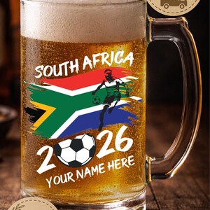 May include: A clear glass beer mug filled with beer and foam. The mug features the South African flag, a soccer player graphic, the numbers "2026", and the text "YOUR NAME HERE".