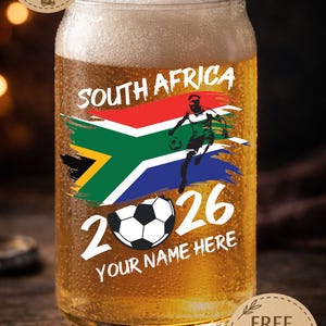 May include: A glass of beer with a design featuring the South African flag, a soccer player, and the text "SOUTH AFRICA 2026". The words "YOUR NAME HERE" are also included. The glass is filled with beer and has condensation.