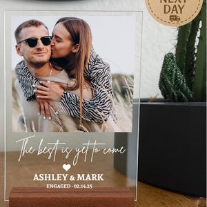May include: Clear acrylic photo frame with a picture of a couple embracing. The frame has a brown wooden base and the text "The best is yet to come" with the names "ASHLEY & MARK" and "ENGAGED - 02.14.25".