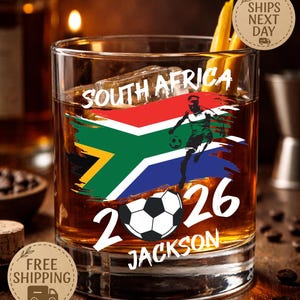 May include: A rocks glass with a South African flag design and a soccer player graphic. The glass features the text "SOUTH AFRICA 2026 JACKSON". The glass contains ice and a dark amber liquid.