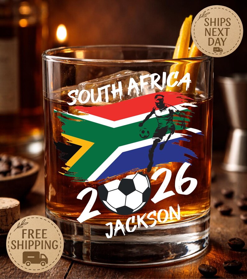 May include: A rocks glass with a South African flag design and a soccer player graphic. The glass features the text "SOUTH AFRICA 2026 JACKSON". The glass contains ice and a dark amber liquid.