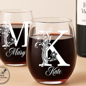 May include: Two stemless wine glasses filled with red wine. Each glass features a large white letter and floral design, with the names "Mary" and "Kate" in script. A bottle of red wine is in the background. The glasses are laser engraved.