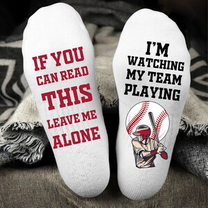 May include: A pair of white socks with red text. One sock says "If you can read this leave me alone". The other sock says "I'm watching my team playing" and has a cartoon baseball player with a bat and ball.