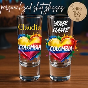 May include: Two personalized shot glasses with a heart-shaped design in the colors of the Colombian flag. One glass has the name "Claudia" in gold, the other says "YOUR NAME". The words "COLOMBIA" are below the heart.