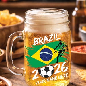 May include: A clear glass beer mug filled with beer and foam. The mug features the word "BRAZIL" in white, a Brazilian flag, a soccer player graphic, a soccer ball, and the number "2026". The text "YOUR NAME HERE" is also visible.