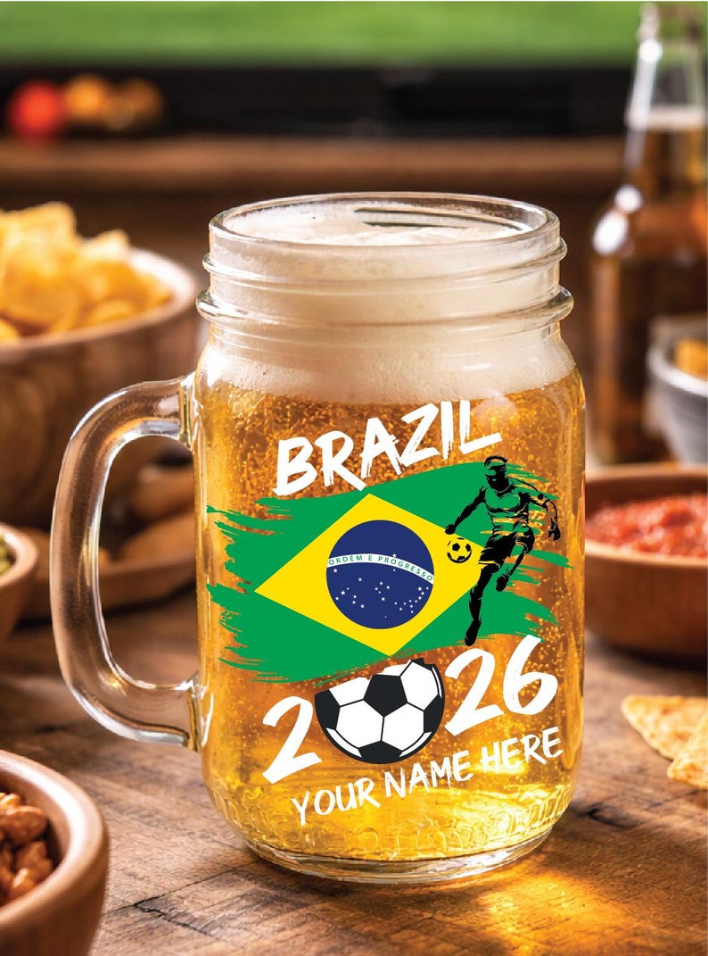 May include: A clear glass beer mug filled with beer and foam. The mug features the word "BRAZIL" in white, a Brazilian flag, a soccer player graphic, a soccer ball, and the number "2026". The text "YOUR NAME HERE" is also visible.