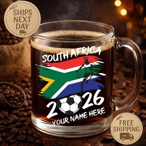 May include: A clear glass coffee mug filled with dark coffee. The mug features a design with the South African flag, a soccer player, a soccer ball, and the text "SOUTH AFRICA 2026 YOUR NAME HERE".